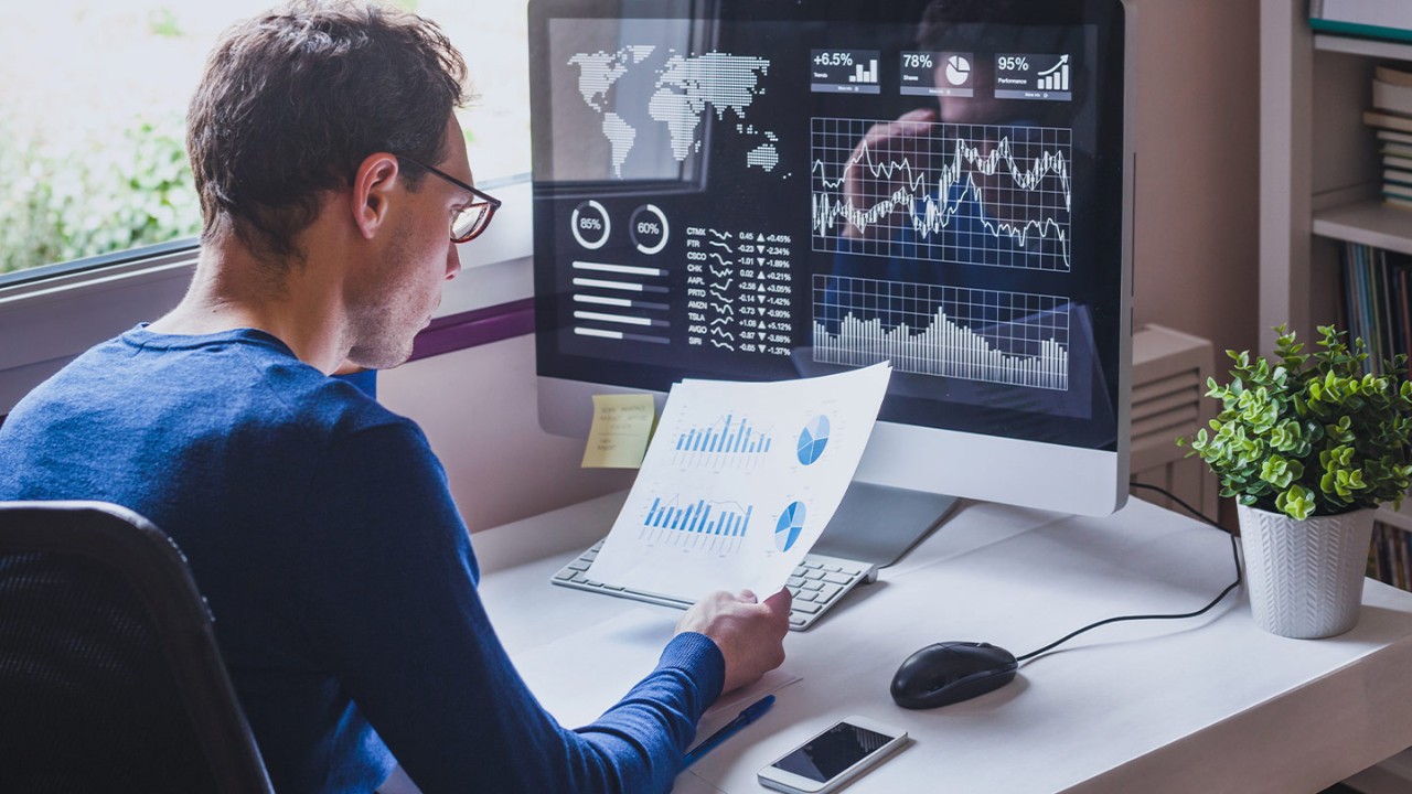 Man sitting at desk looking at data analytic reports on paper and on his desktop monitor in front of him.