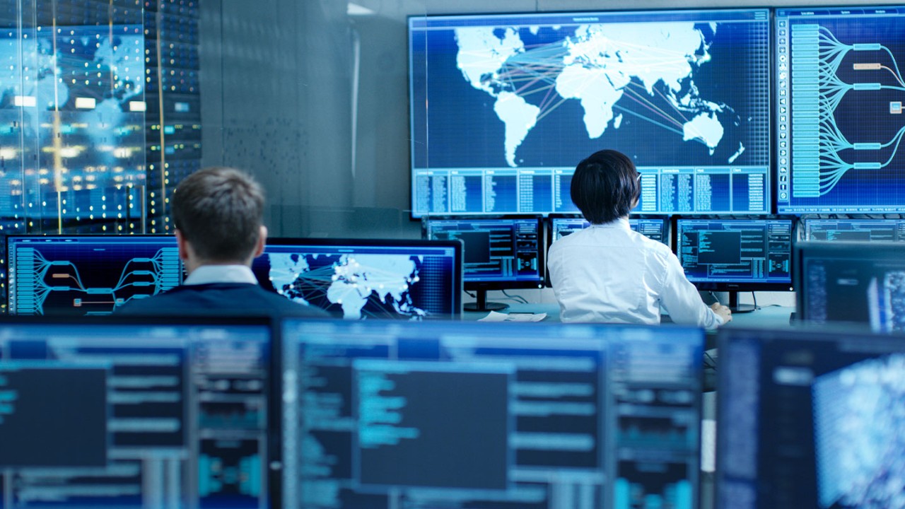 Two men sitting at desks in a mission control center looking at monitors surrounding them.