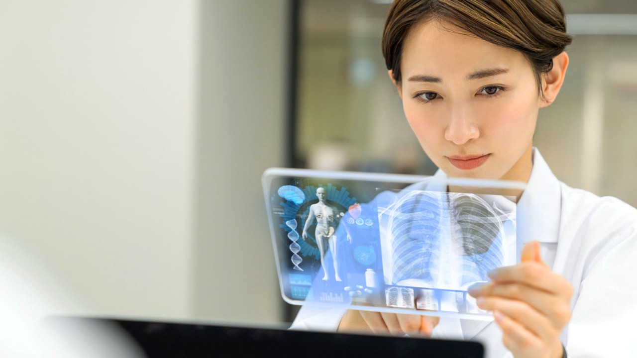 Woman in lab coat looking at a clear tablet showing patient diagnostics while she types information into a laptop.