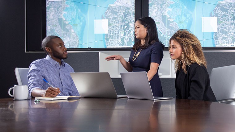 Woman standing and presenting to a man and woman sitting at a conference table with laptops in front of them. 