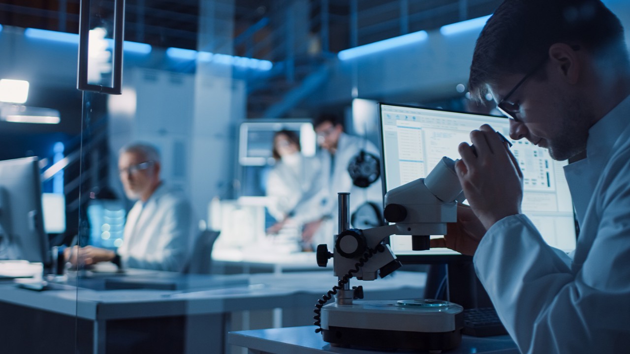 Scientist at a desk looking through a microscope with data on a monitor next to him surrounded by other scientists working at their desks.