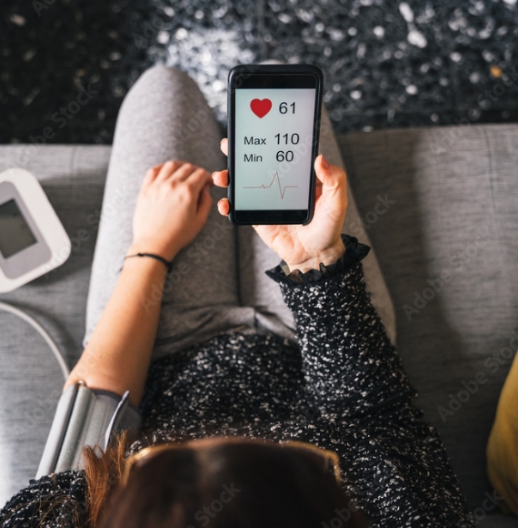 Woman sitting on a couch with a heart monitor cuff on her arm while looking at the connected app showing results on her phone.